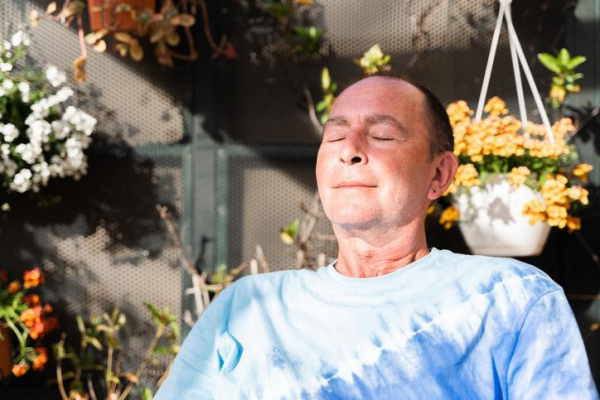 An older man calmly meditatating while seated in a sunny spot with eyes closed and a slight smile; hanging flowers in the background